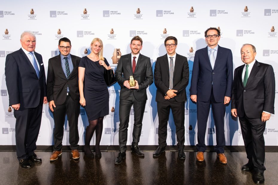 In front of a photo wall, representatives of Cubicure GmbH in formal attire stand together with representatives of the Houskapreis. Dr. Robert Gmeiner holds the Houskapreis 2019 awarded to the company.