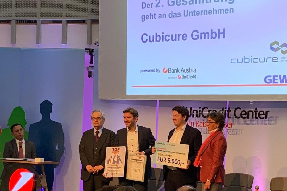 Representatives of Cubicure GmbH accept the 2nd prize of the JUNGUNTERNEHMER competition 2019 on a stage. They hold certificates and a check for the prize money. With them on the stage are two award representatives.
