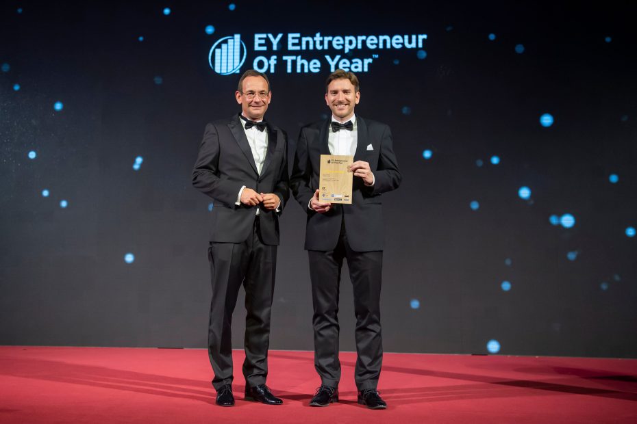 The CEO of Cubicure GmbH stands on a stage with a representative of Ernst & Young. In his hands, he holds an award honoring him as an Entrepreneur of the Year winner.