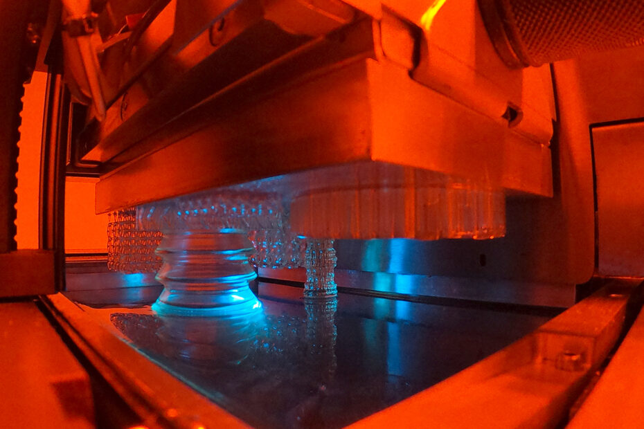 Ein Blick in das Innere von Cubicures 3D-Drucker Caligma 200. Durchsichtige Komponenten sind gerade im Druckprozess inbegriffen. Die Szene ist in oranges und blaues Licht getaucht.