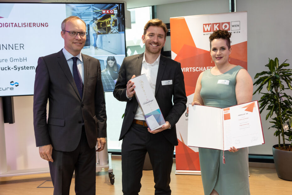 Three people stand in front of a screen and a rollup bearing the logo of the Vienna Chamber of Commerce. From left to right: Alexander Biach of WK Wien; Dr. Robert Gmeiner, CEO & CTO of Cubicure GmbH, holding a champagne bottle; Kathrin Wallner of Cubicure GmbH, with the award certificate.