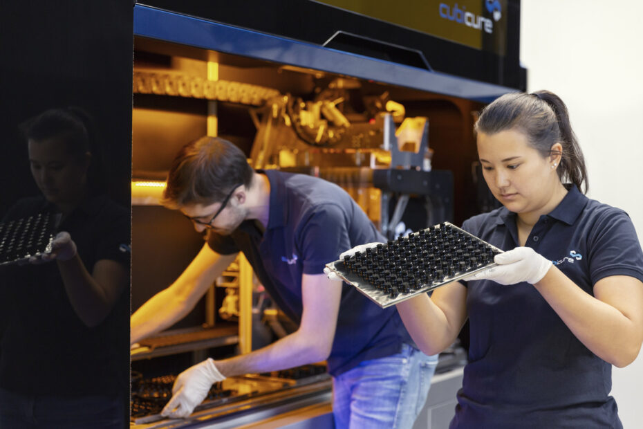 Zwei Personen arbeiten an einem großen 3D-Druck-System. Beide tragen Cubicure T-Shirts. Der Mann im Hintergrund hebt eine Bauplattform aus der Maschine. Im Vordergrund kontrolliert eine Frau die Qualität der gedruckten Bauteile.