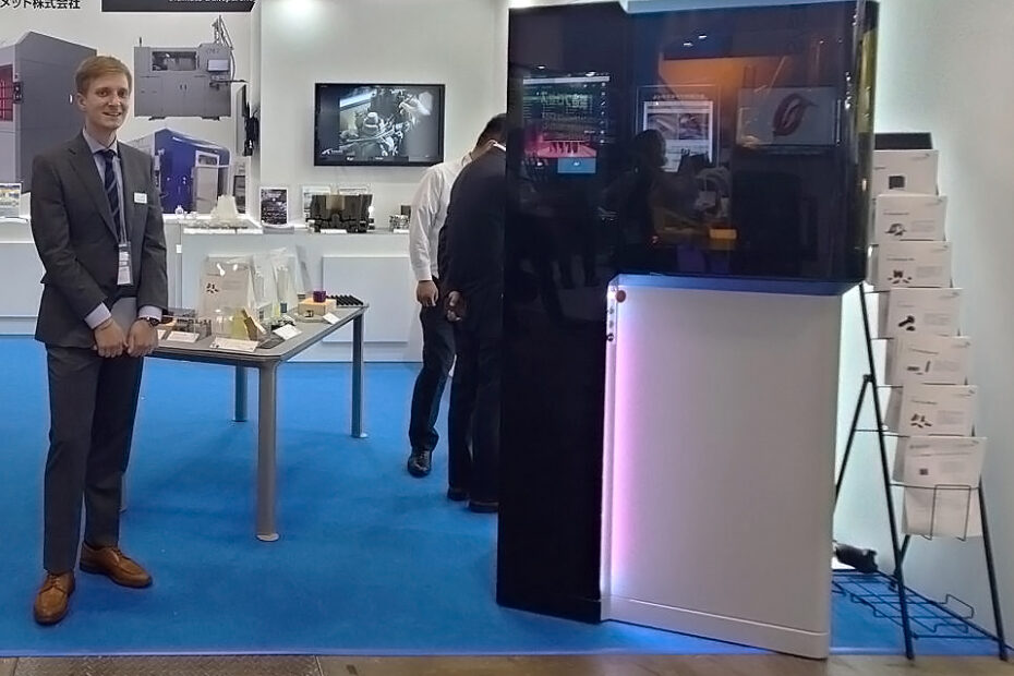 A trade show booth at the exhibition Manufacturing World Japan. On the left side, a white man in a suit stands smiling. He is a Cubicure sales representative. On the right side, a sleek black machine; a Caligma 3D printer.